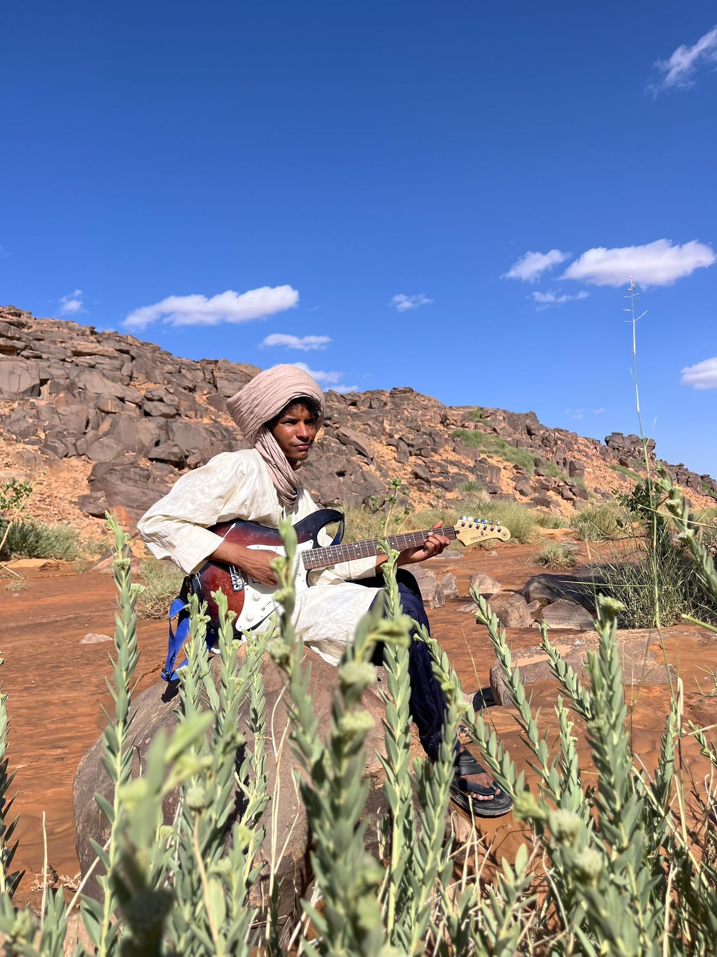 Khily Ag Osman con tagelmust, tocando la guitarra eléctrica entre plantas del desierto
