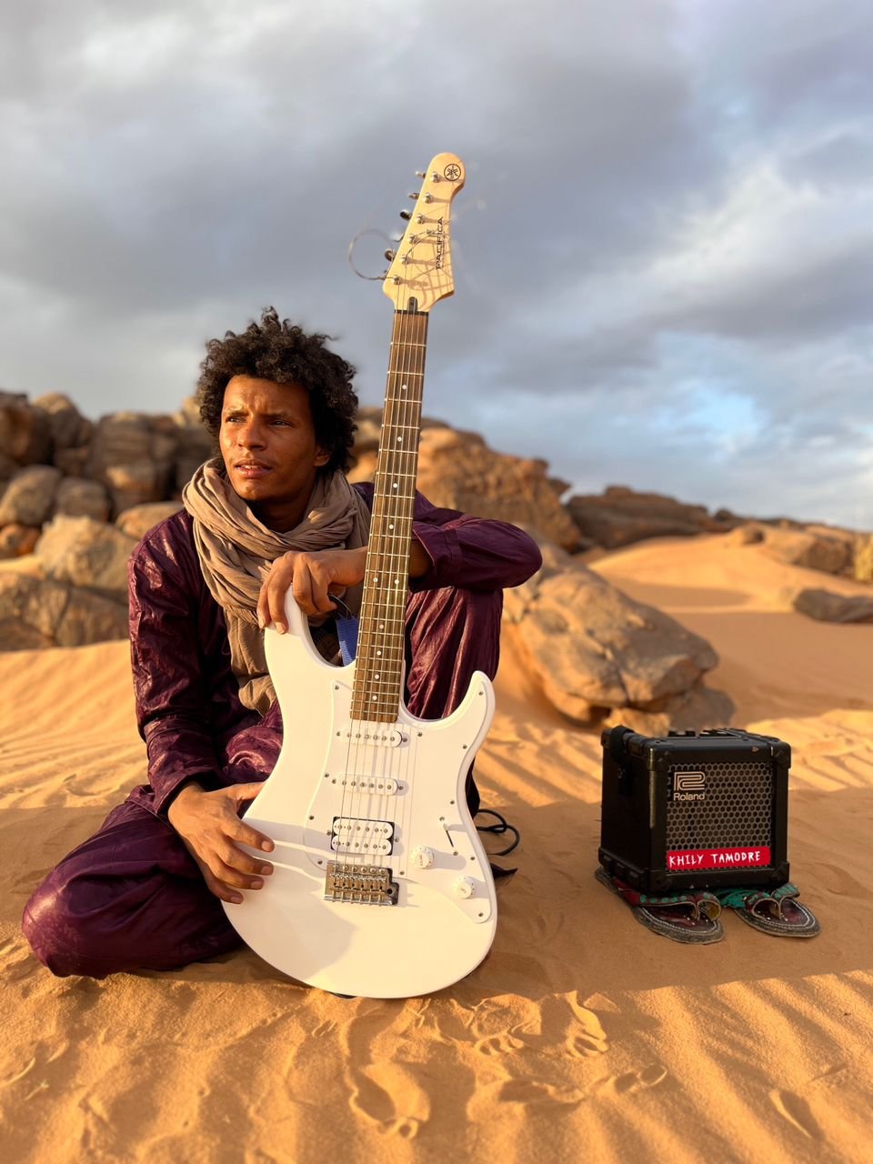 Khily Ag Osman sentado en la arena con su guitarra eléctrica blanca y su amplificador, entre las rocas de Djanet